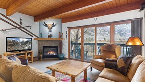 A cozy living room with a fireplace, a television, and a large window overlooking a deck. - Riverside Condos B101 by Avantstay Condo Close To Downtown, Town Park & Ski Lift #8 (Telluride)
