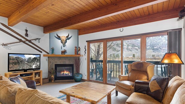 A cozy living room with a fireplace, a television, and a large window overlooking a deck. - Riverside Condos B101 by Avantstay Condo Close To Downtown, Town Park & Ski Lift #8 (Telluride)