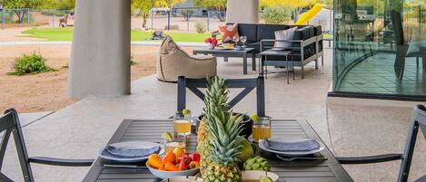 An outdoor dining area with a table set for two, featuring a tropical fruit arrangement and a glass of beverage.