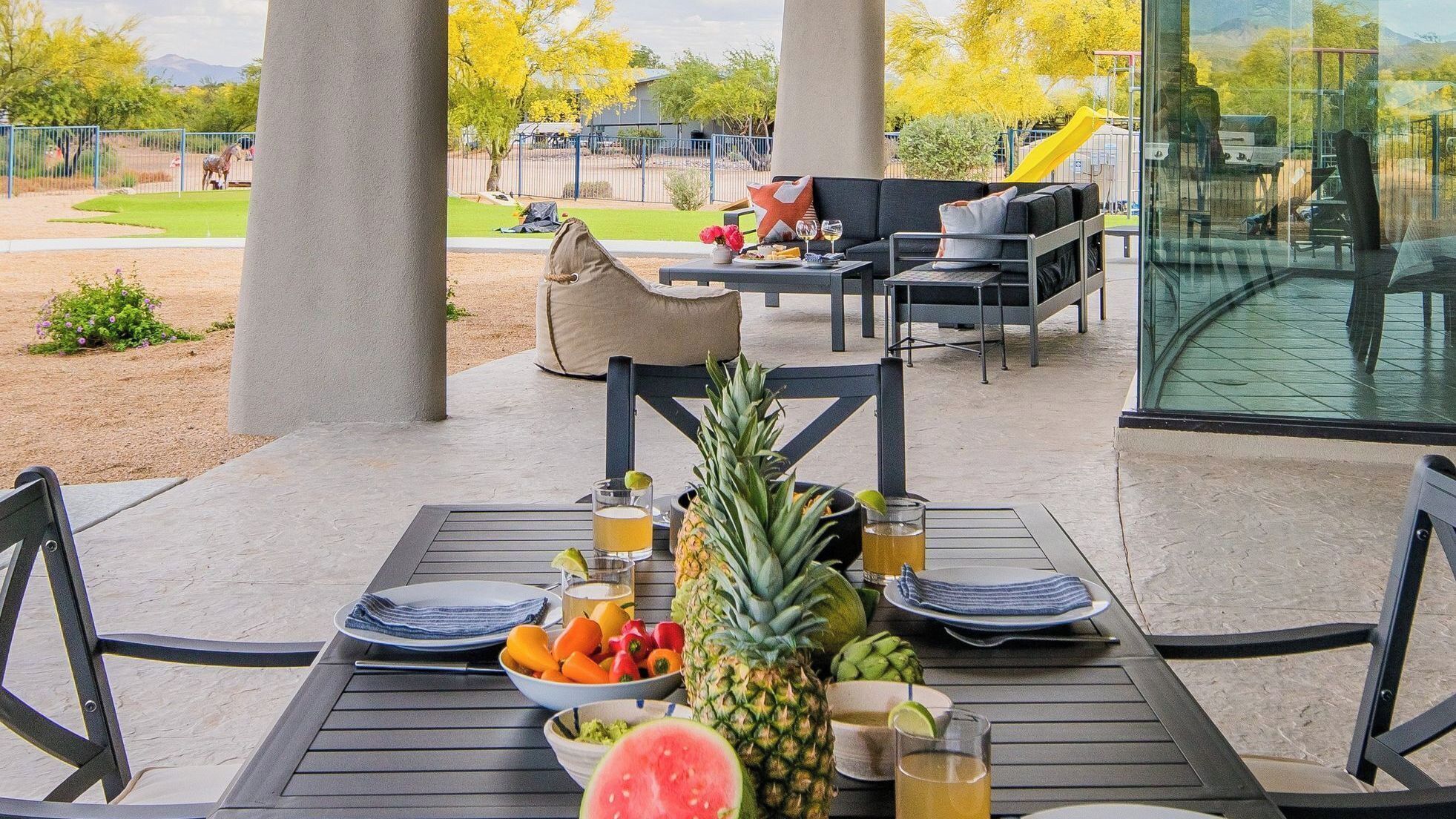 An outdoor dining area with a table set for two, surrounded by chairs, a sofa, and a glass enclosure.