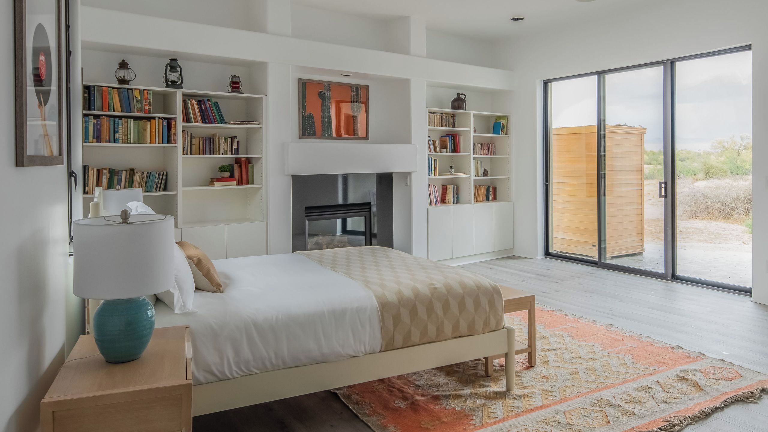 A modern bedroom with a bed, bookshelves, a fireplace, and a large sliding glass door to the outside.
