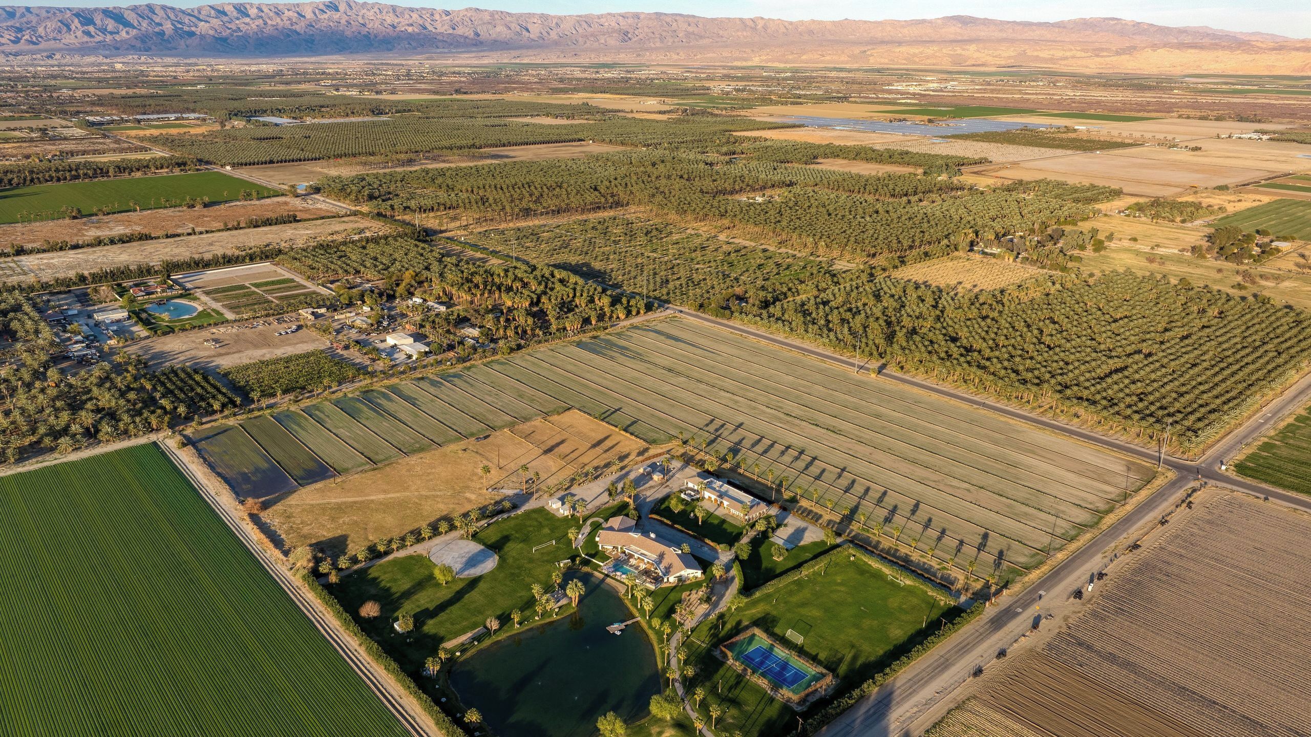 Luftaufnahme einer weiten Agrarlandschaft mit Feldern, einem Golfplatz und einem Wohngebiet mit Häusern und einem Schwimmbad.
