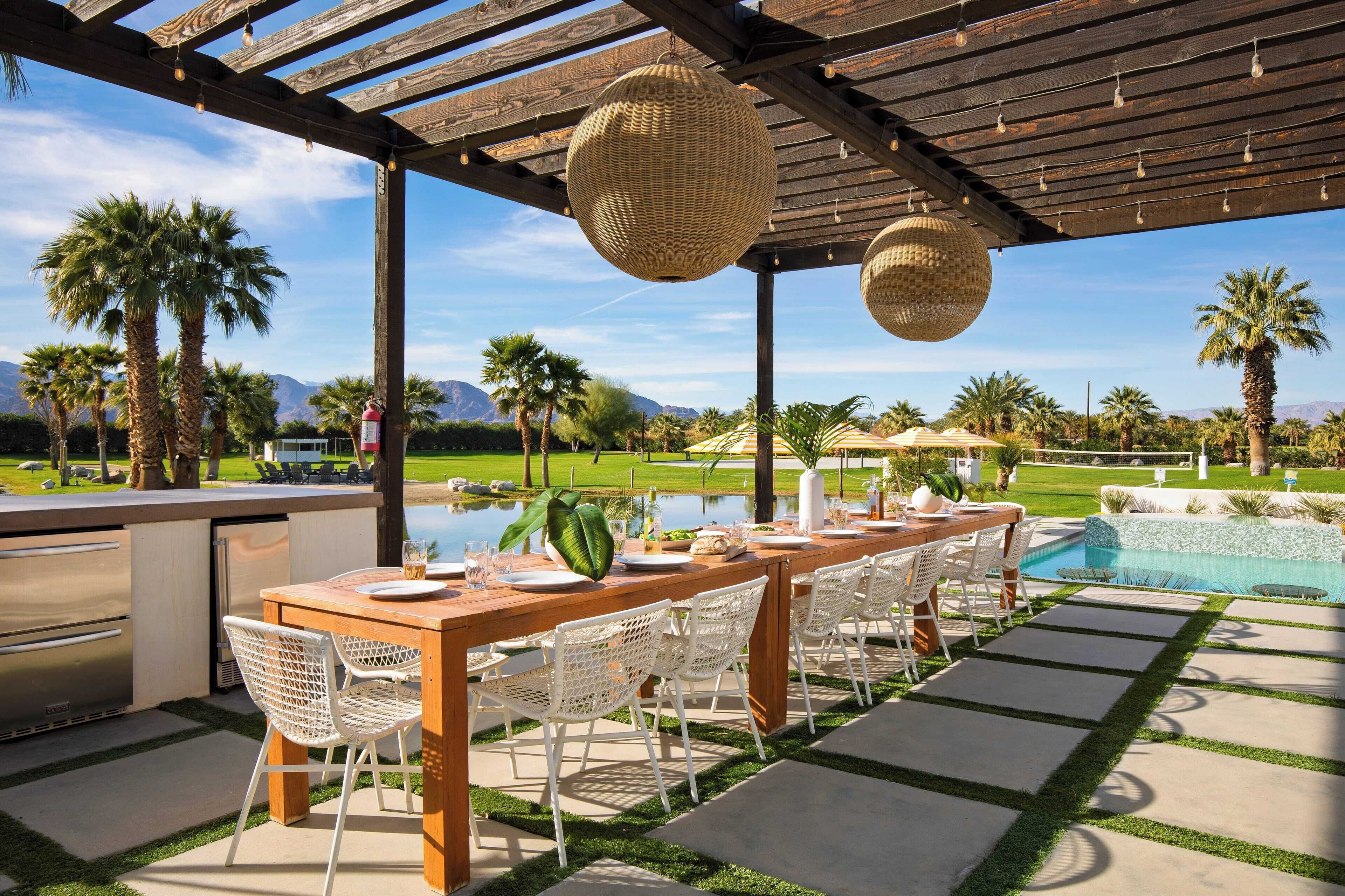 Un espace de restauration en plein air avec une table en bois et des chaises blanches, une piscine, des palmiers et des montagnes en arrière-plan.