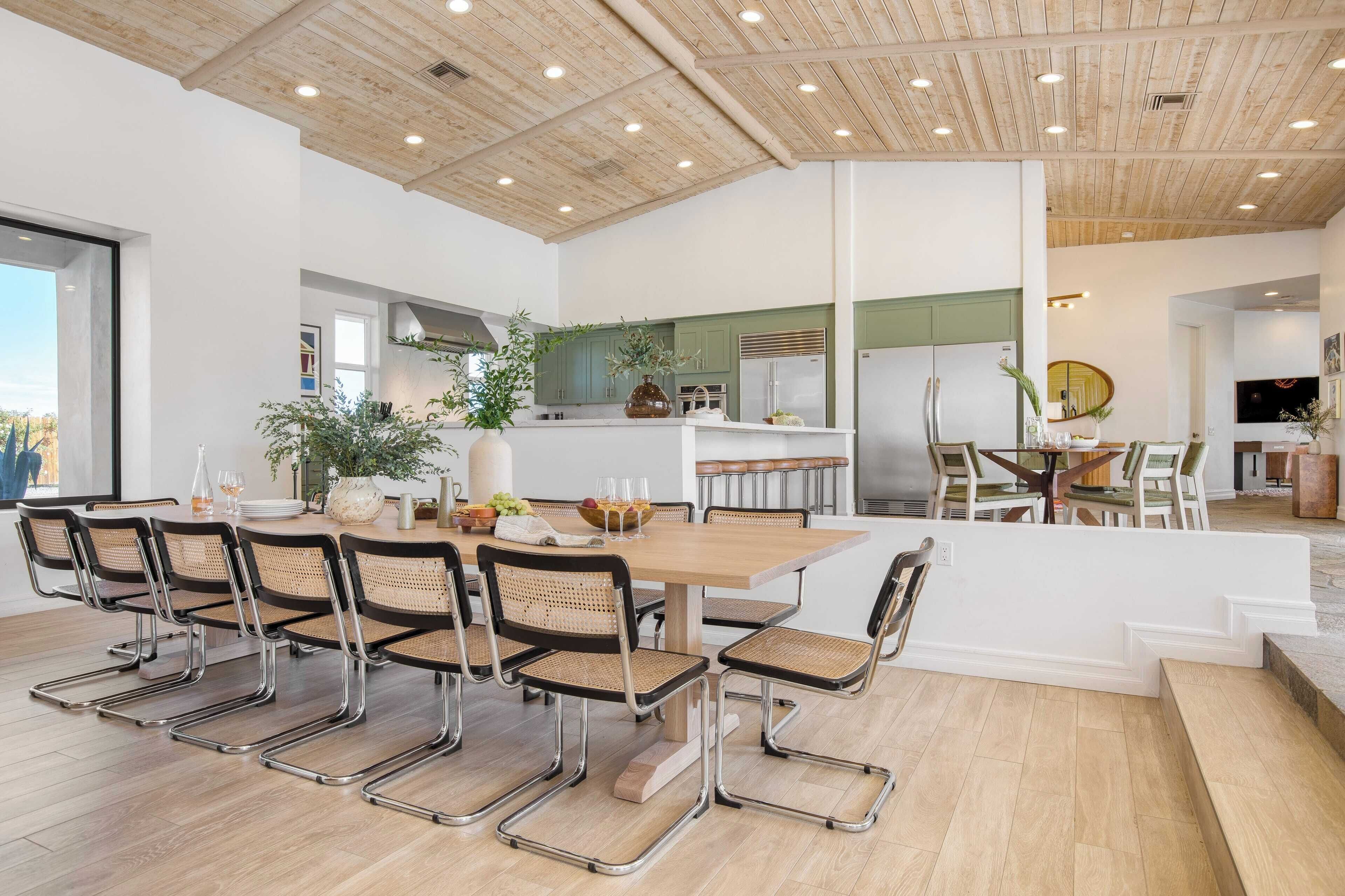 Un grand espace à manger avec une table en bois et des chaises en métal, une cuisine en arrière-plan et une vue sur l’extérieur.