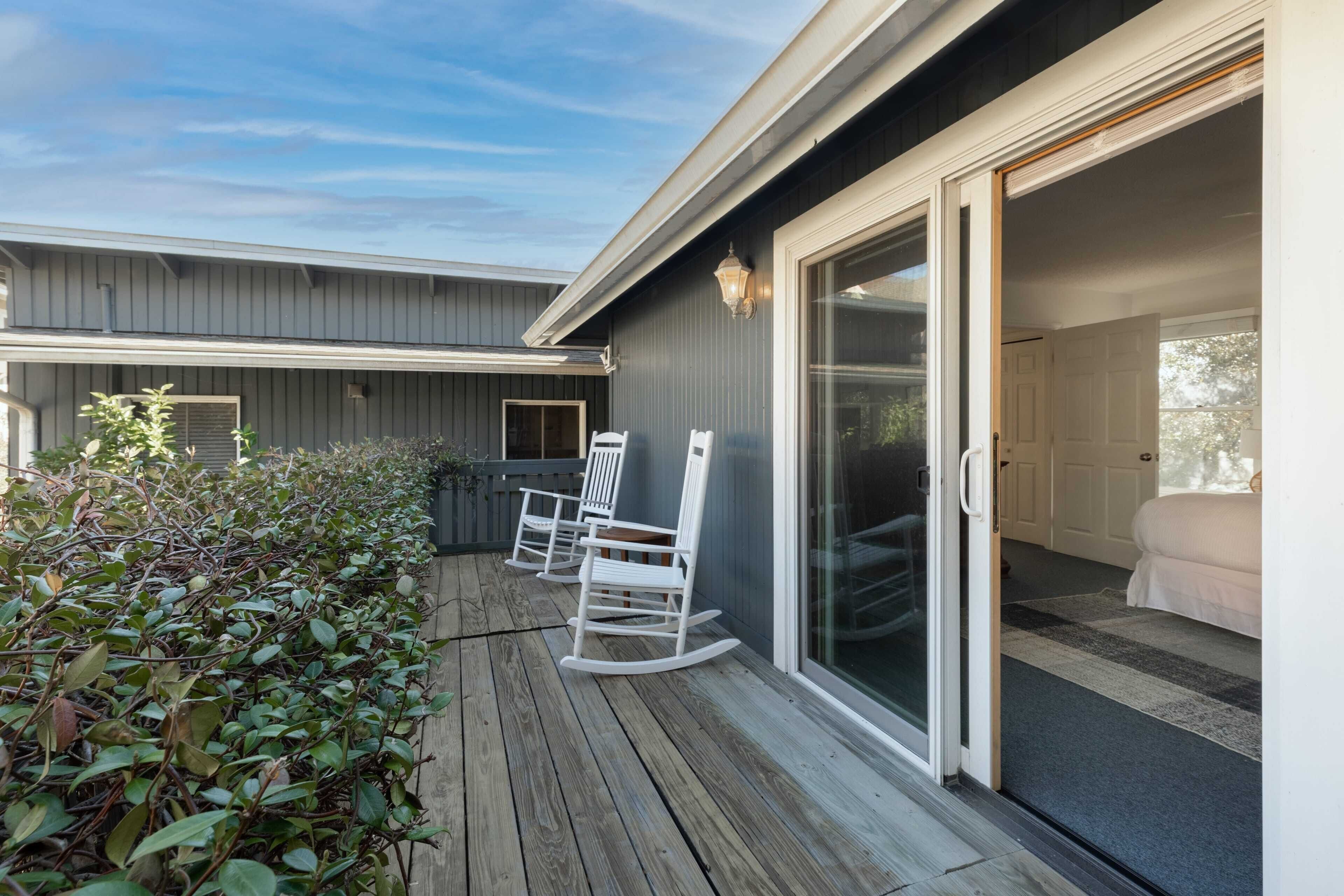 A wooden deck with white rocking chairs, a sliding glass door leading to a bedroom, and a small outdoor area with plants.