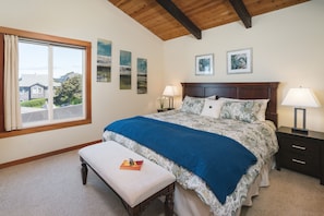 A bedroom with a large bed, a bench, a nightstand, and a window with a view of houses. - Pacific House by Avantstay Bright Airy Home w/ Direct Access to Cannon Beach (Cannon Beach)
