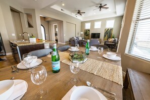 Un coin repas avec une table en bois dressée pour quatre personnes, comprenant des verres à vin, des assiettes et des couverts. On y trouve deux ventilateurs de plafond, un coin cuisine avec un évier, et un salon avec un canapé et un téléviseur.