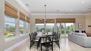 Un comedor amplio con una mesa grande y sillas, ventanas grandes con persianas, una araña y vistas al exterior.