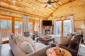 L’intérieur chaleureux d’un chalet avec un plafond en bois, une cheminée en pierre, un téléviseur et une grande fenêtre donnant sur une forêt.