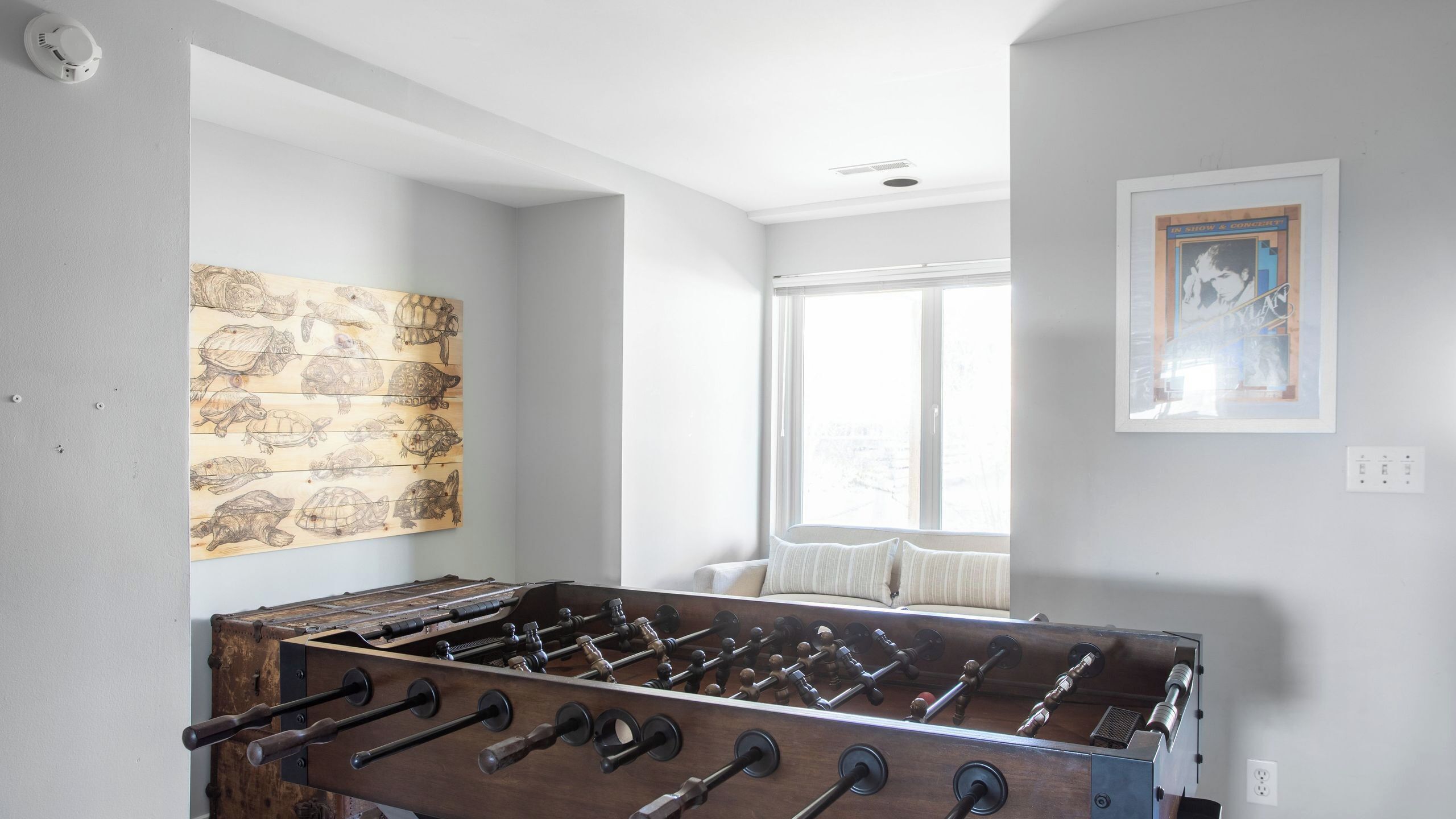 A room with a foosball table, a wall art of turtles, a window with curtains, and a framed picture on the wall.