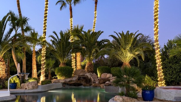 A pool area with palm trees and string lights, surrounded by rocks and plants. - Preston by Avantstay 6BR Desert Oasis w/ Basketball Court, Pool & Hot Tub (Scottsdale)
