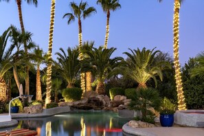 Un área de piscina con palmeras y luces colgantes, rodeada de rocas y plantas.
