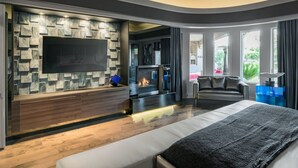 A modern bedroom with a large bed, a fireplace, a TV mounted on a stone wall, and a view of the outdoors. - Preston by Avantstay 6BR Desert Oasis w/ Basketball Court, Pool & Hot Tub (Scottsdale)