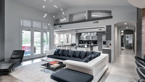 A modern living room with a white sofa and black cushions, a black coffee table with books, and a large window with a view of greenery. - Preston by Avantstay 6BR Desert Oasis w/ Basketball Court, Pool & Hot Tub (Scottsdale)