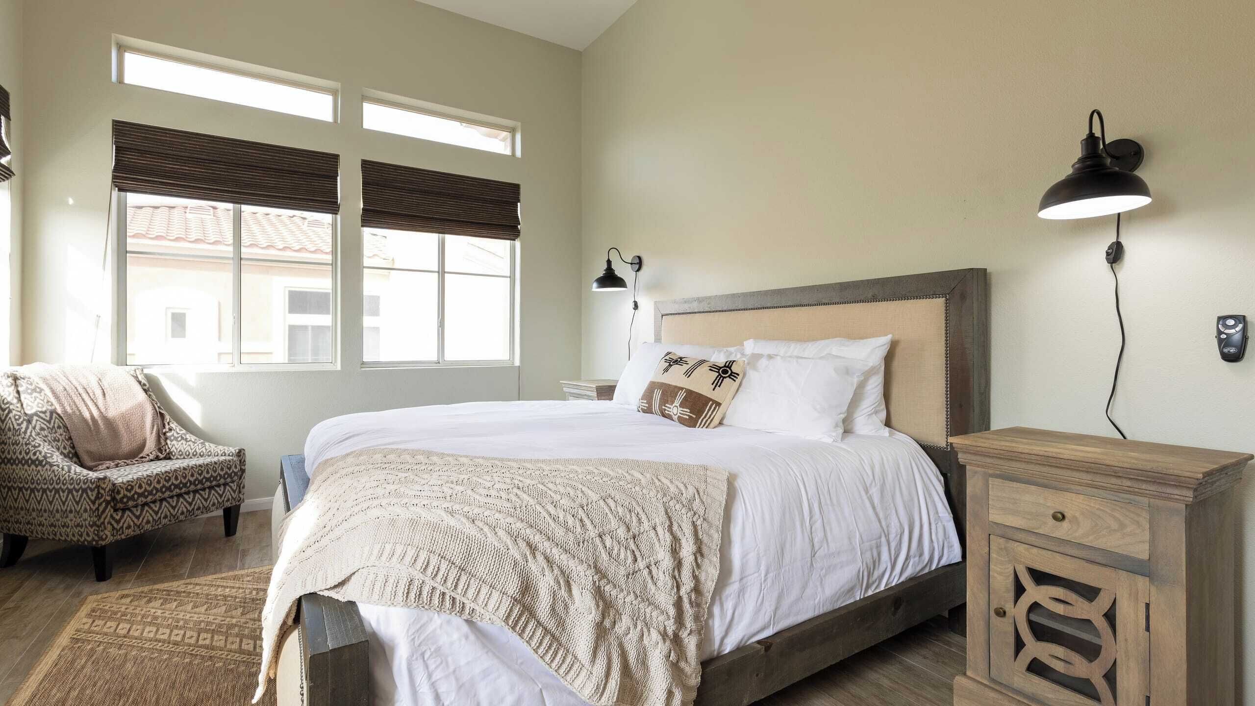 A bedroom with a large bed, a chair, a nightstand, a lamp, and a window with blinds.