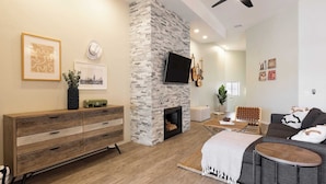 A modern living room with a stone fireplace, a flat-screen TV, and a sofa. - Eucalyptus by Avantstay Stunning Scottsdale Home w/ Communal Pool & Hot Tub (Scottsdale)