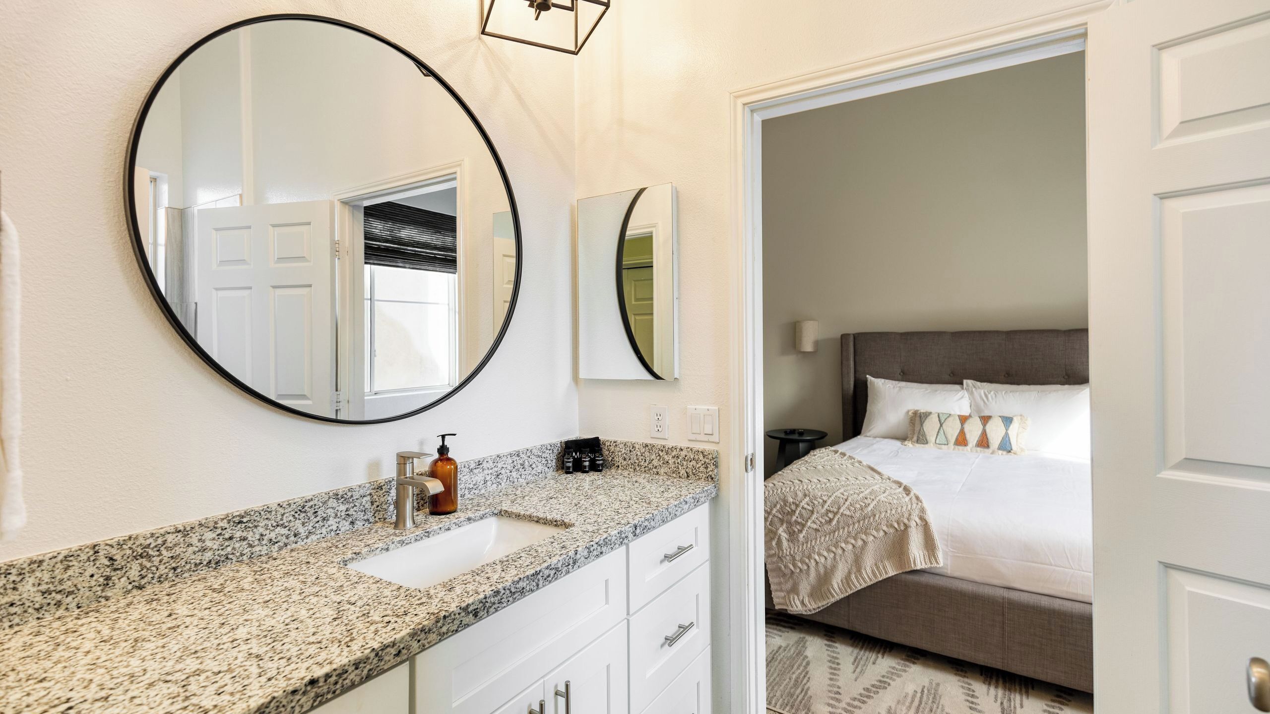 A bathroom with a granite countertop, a round mirror, and a bed with a gray headboard in the adjacent room.