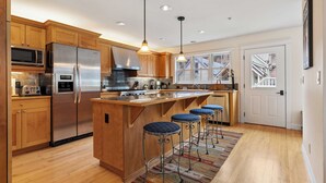 A modern kitchen with wooden cabinets, stainless steel appliances, a central island with bar stools, and a view of the outside through large windows. - Legacy House by Avantstay Beautiful 4bdr Home in Great Location w/ Hot Tub (Telluride)