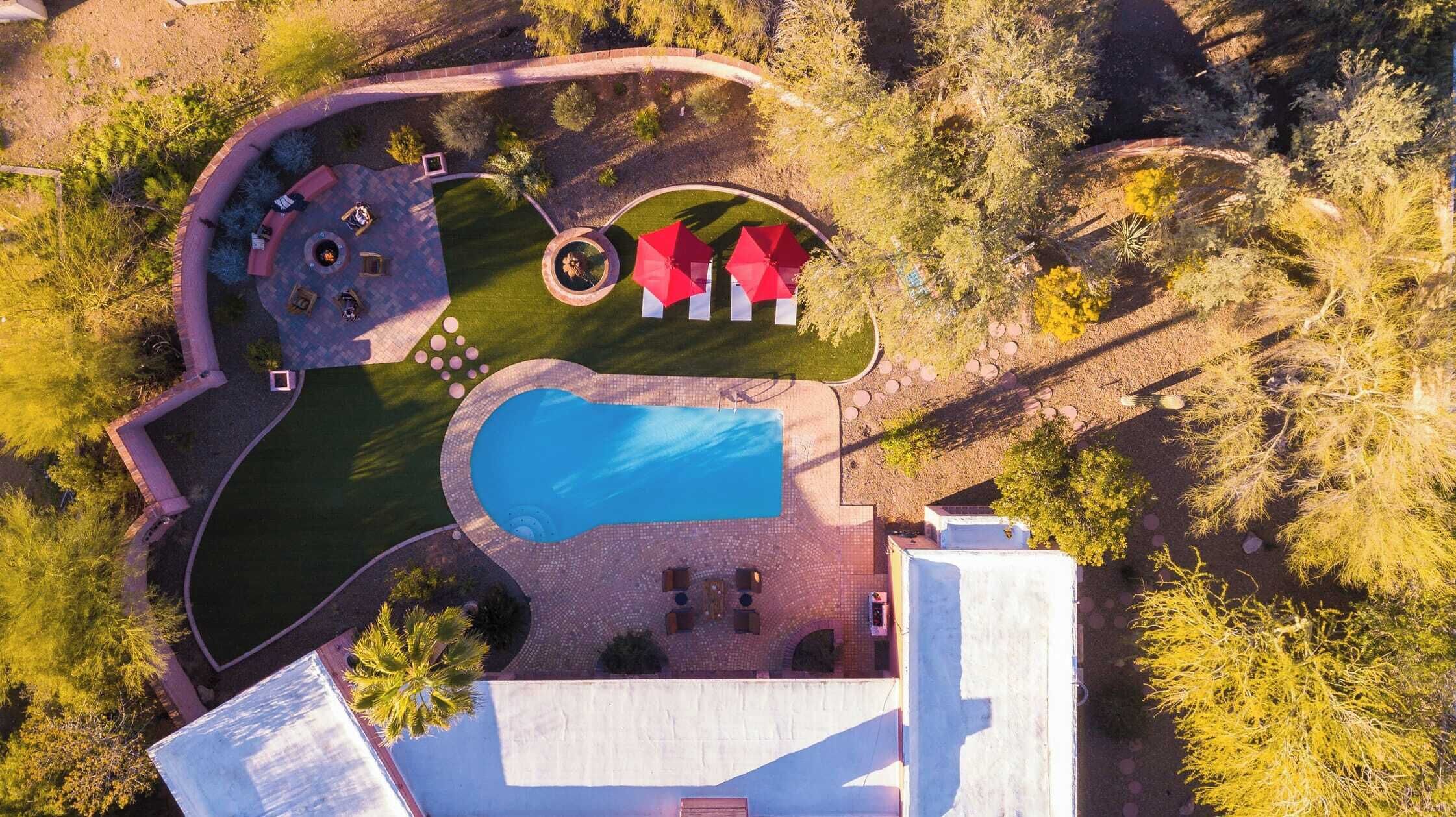 Eine Vogelperspektive eines angelegten Anwesens mit Schwimmbad, Terrassenbereichen und einem kreisförmigen Garten mit roten Sonnenschirmen.