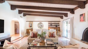 A spacious living room with a fireplace, a coffee table with books, and a large TV. There are two sofas with colorful cushions and a large window providing natural light.