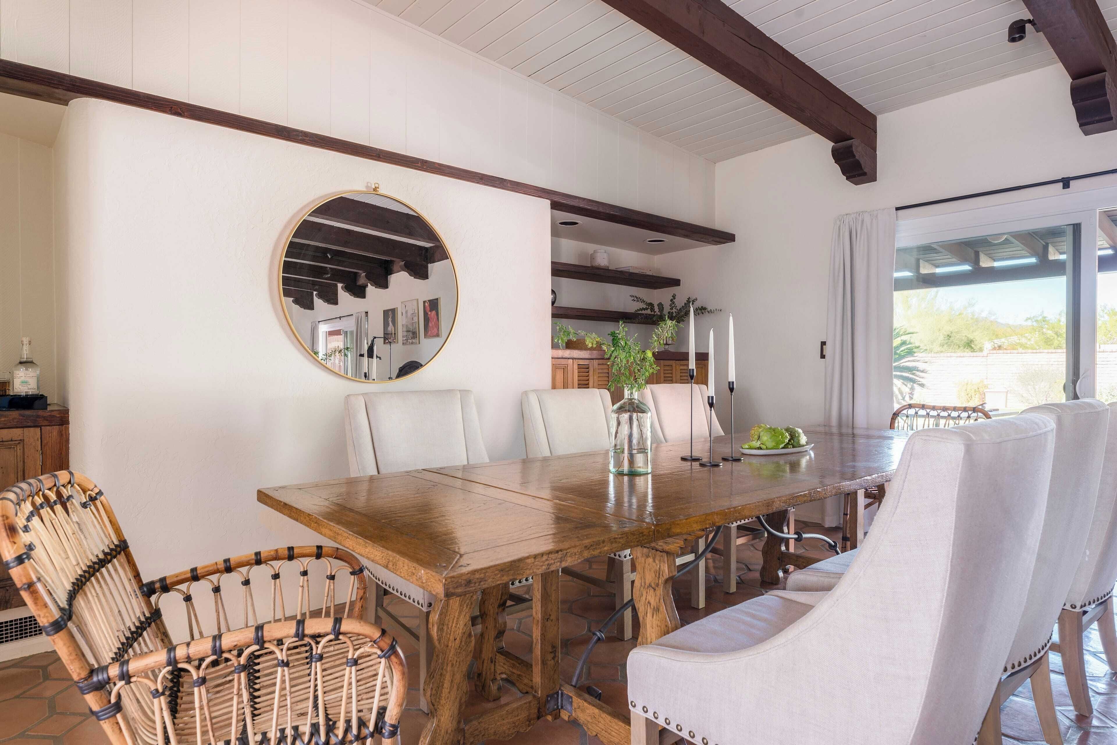 A dining area with a wooden table, white upholstered chairs, a round mirror, and a view of the outdoors through a window.