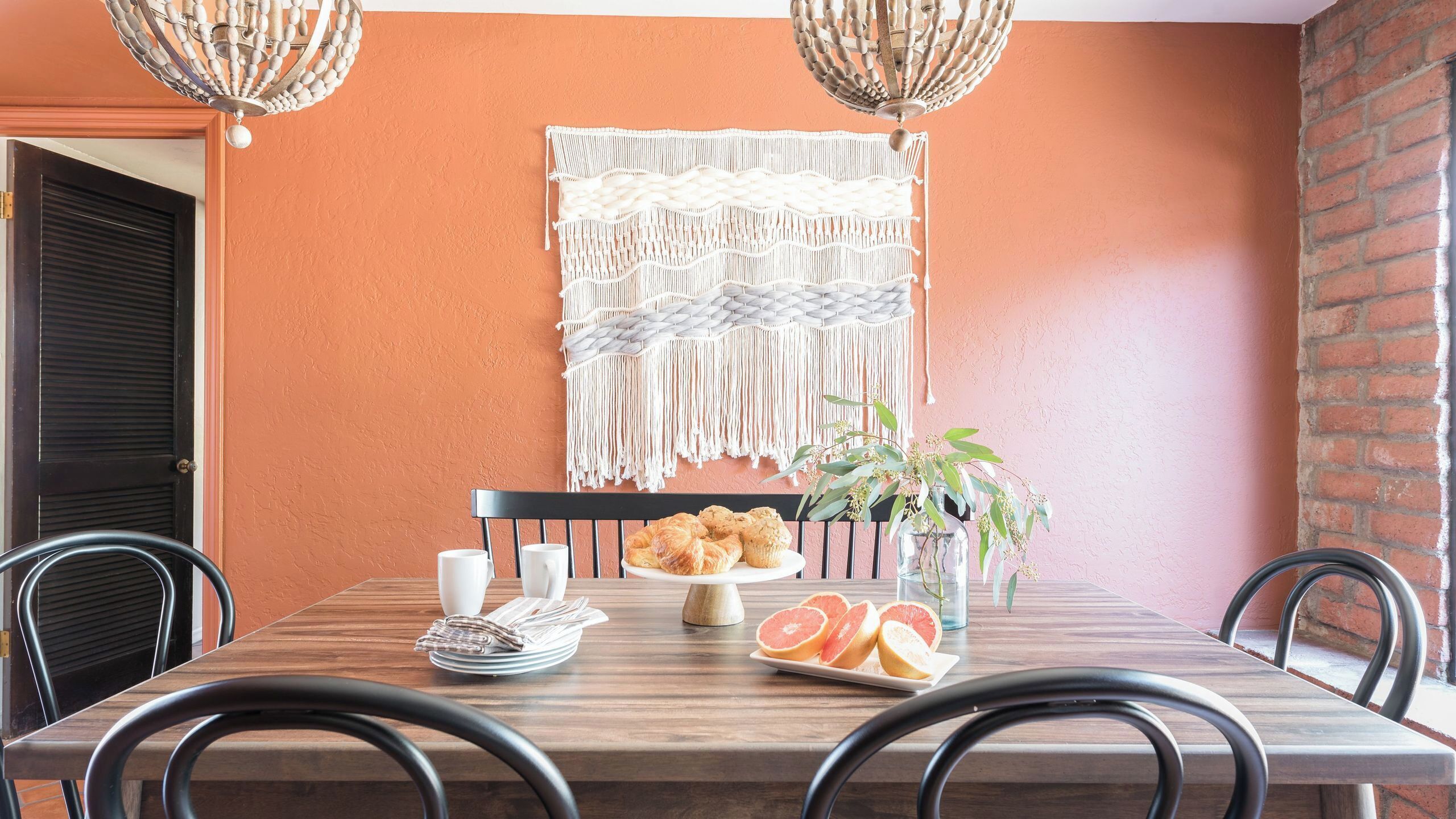 A dining table with a plate of croissants, a glass vase with a plant, and a plate of sliced fruits.