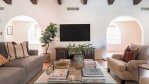 A living room with a sofa, armchair, coffee table, and a TV. There are books and a plant on the table.