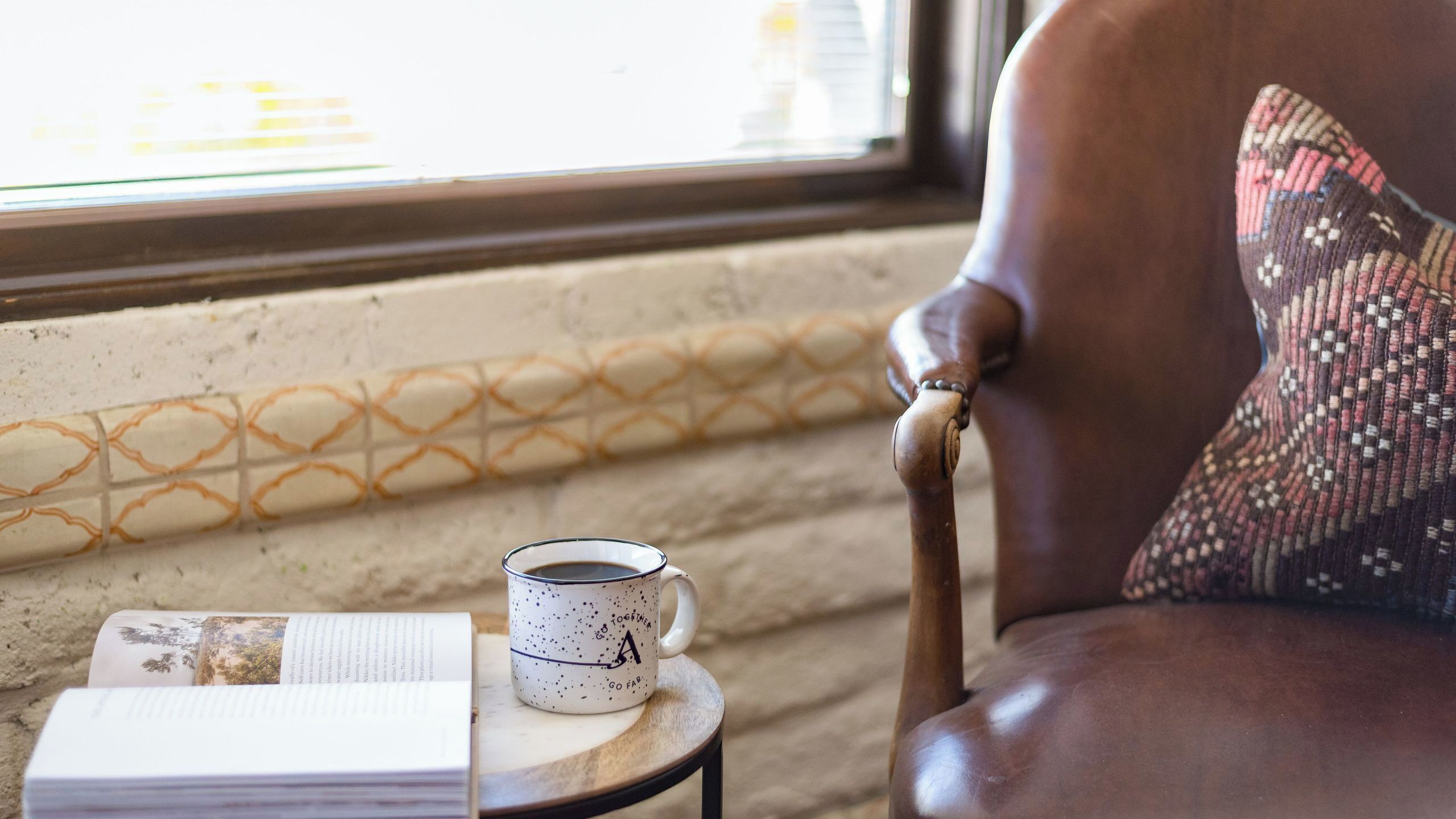 A chair with a patterned cushion, a cup with text, and a book on a small table by a window.