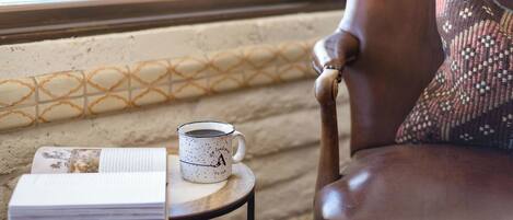 A chair with a patterned cushion, a cup with text, and a book on a small table by a window.