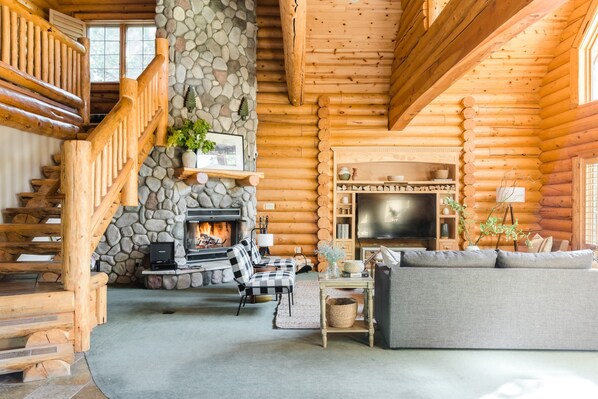 A log cabin interior with a stone fireplace, wooden staircase, and a cozy seating area. - Grizzly by Avantstay Rustic Big Bear Cabin w/ Hot Tub & Pool Table (Big Bear Lake)