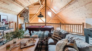 Ett mysigt rum i loftstil med ett träpanelat tak, ett biljardbord, en soffa och ett matbord med en rutig duk.