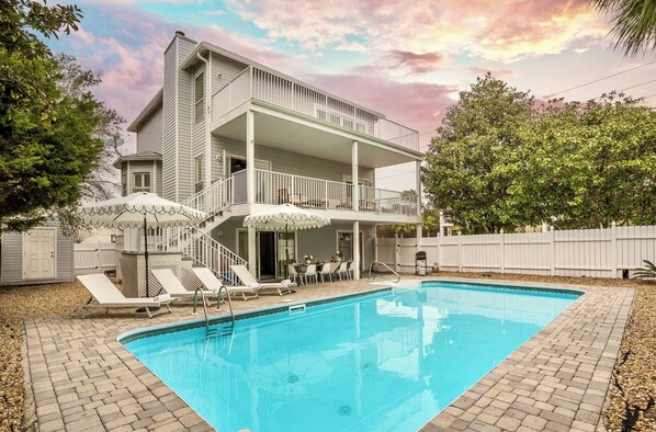 A two-story house with a swimming pool, a covered patio with umbrellas, and a fenced backyard.