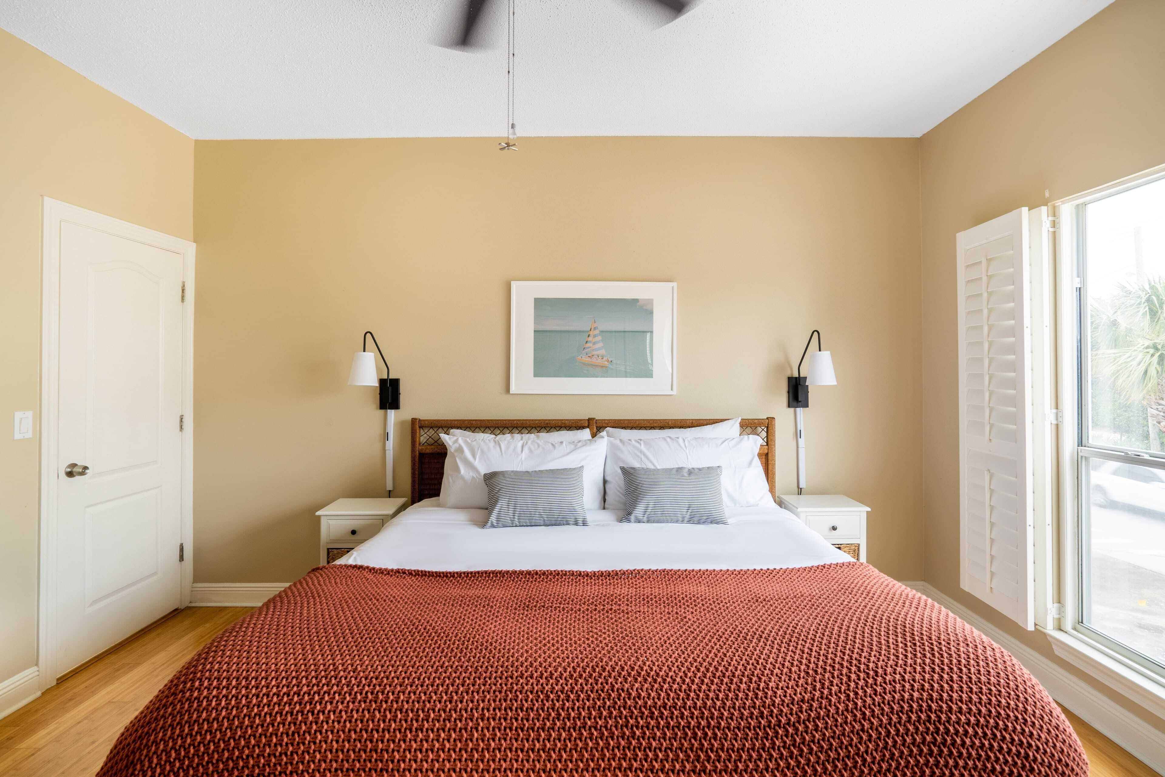 Un dormitorio con cama, ventilador de techo, ventana con contraventanas y un cuadro en la pared.