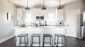 A modern kitchen with white cabinets, a central island, bar stools, and pendant lighting. - Rhett by Avantstay Stylish Southern Style Townhouse w/ Gorgeous Design (Nashville)