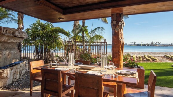 An outdoor dining area with a view of the beach and ocean, wooden furniture, and a stone wall.