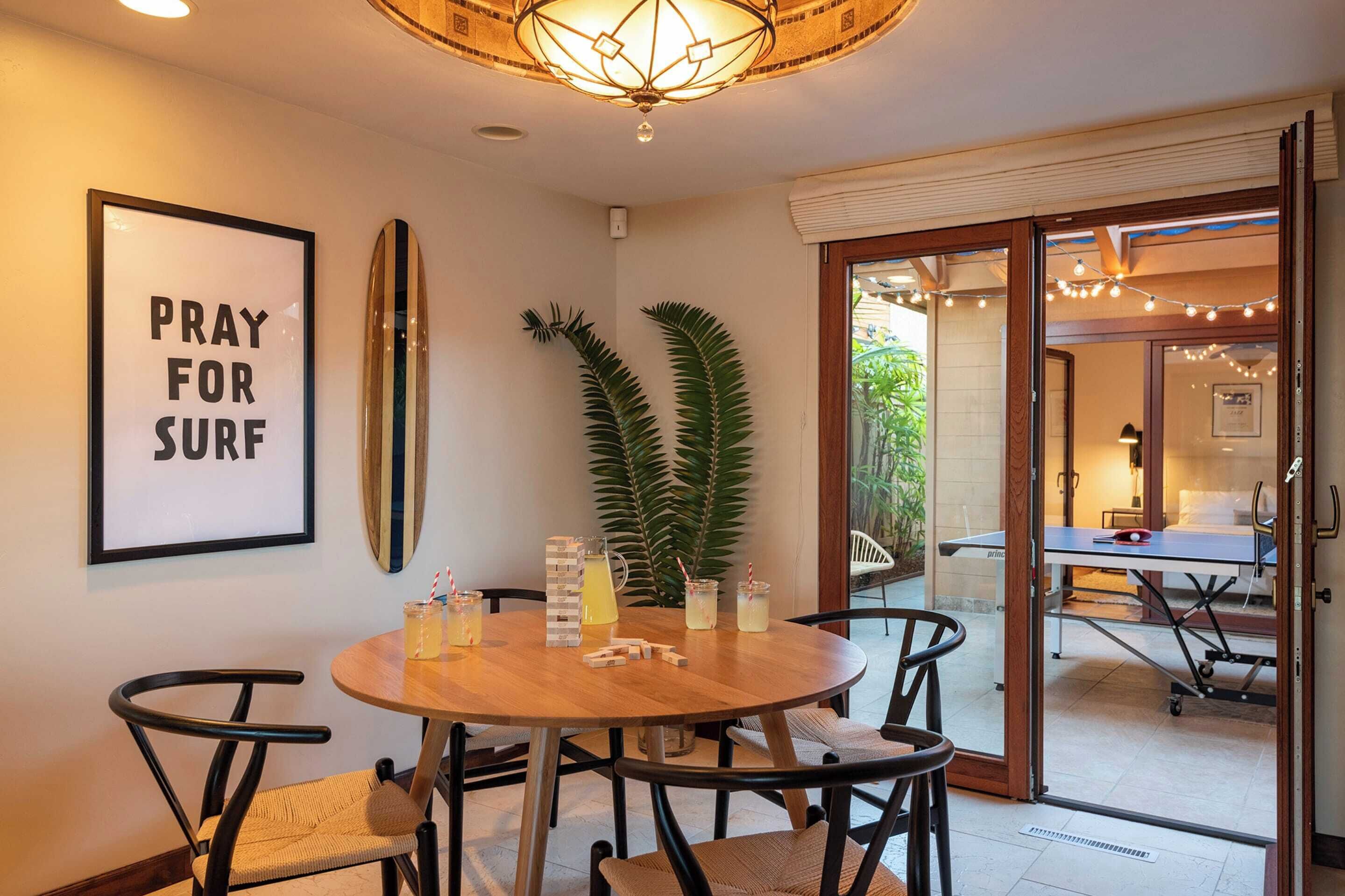 A dining area with a round wooden table, black chairs, a framed sign, a surfboard, and a potted plant.