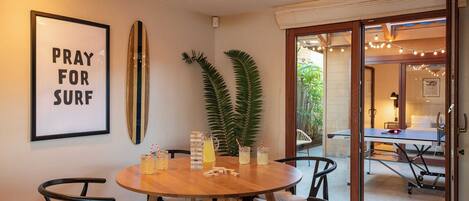 A dining area with a round wooden table, black chairs, a framed sign, a surfboard, and a potted plant.