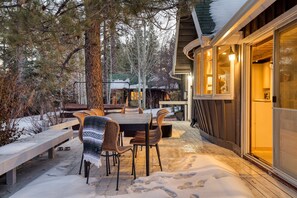 A wooden deck with a table and chairs, surrounded by snow and trees.