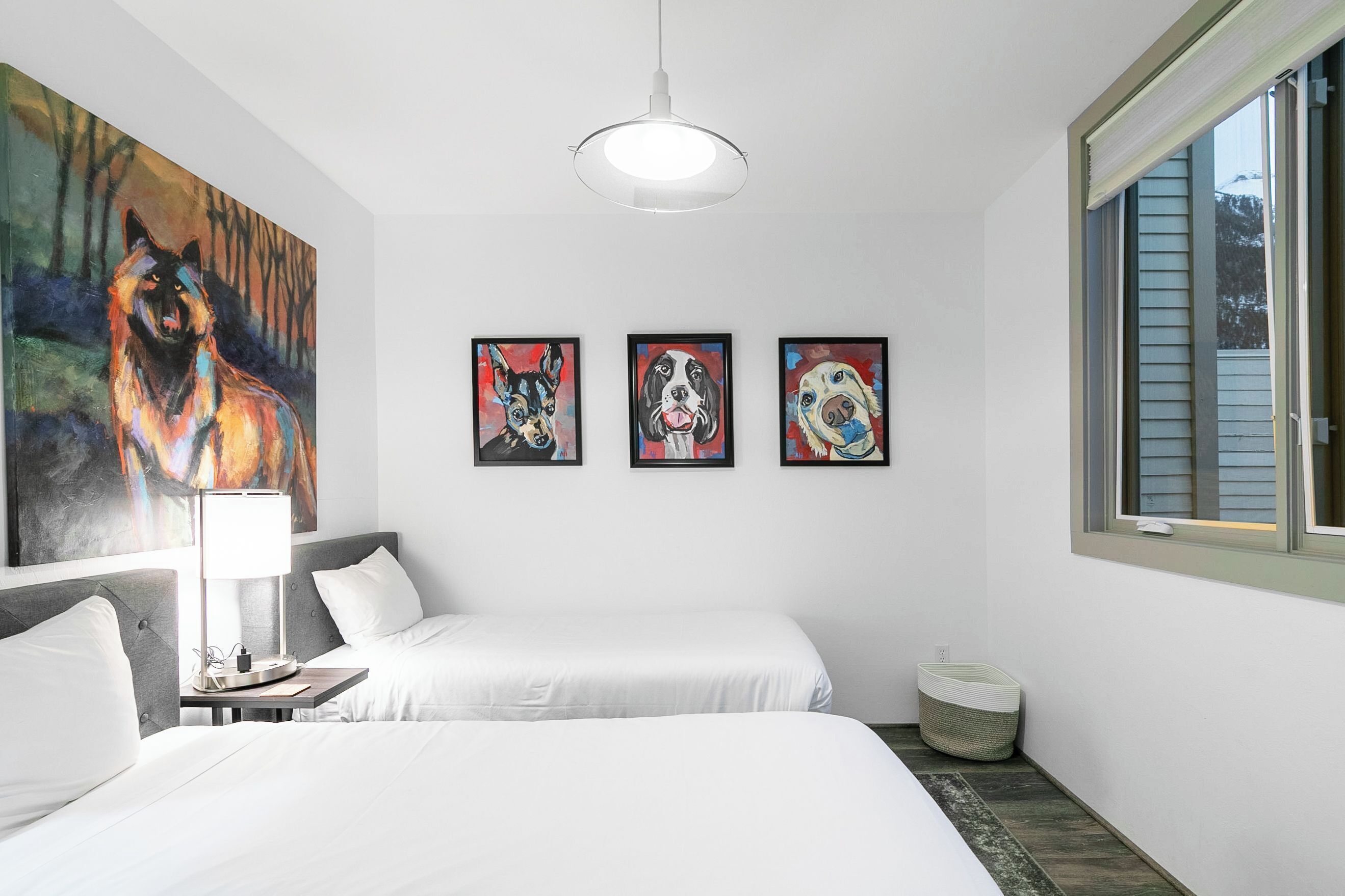 Habitación de hotel con dos camas, un cuadro de un lobo, tres retratos enmarcados de perros, una lámpara y una ventana con persianas.