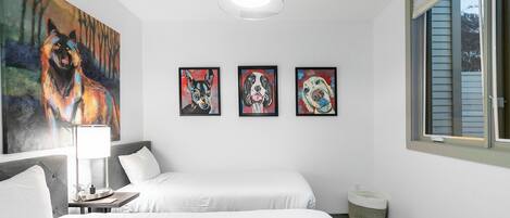 Habitación de hotel con dos camas, un cuadro de un lobo, tres retratos enmarcados de perros, una lámpara y una ventana con persianas.