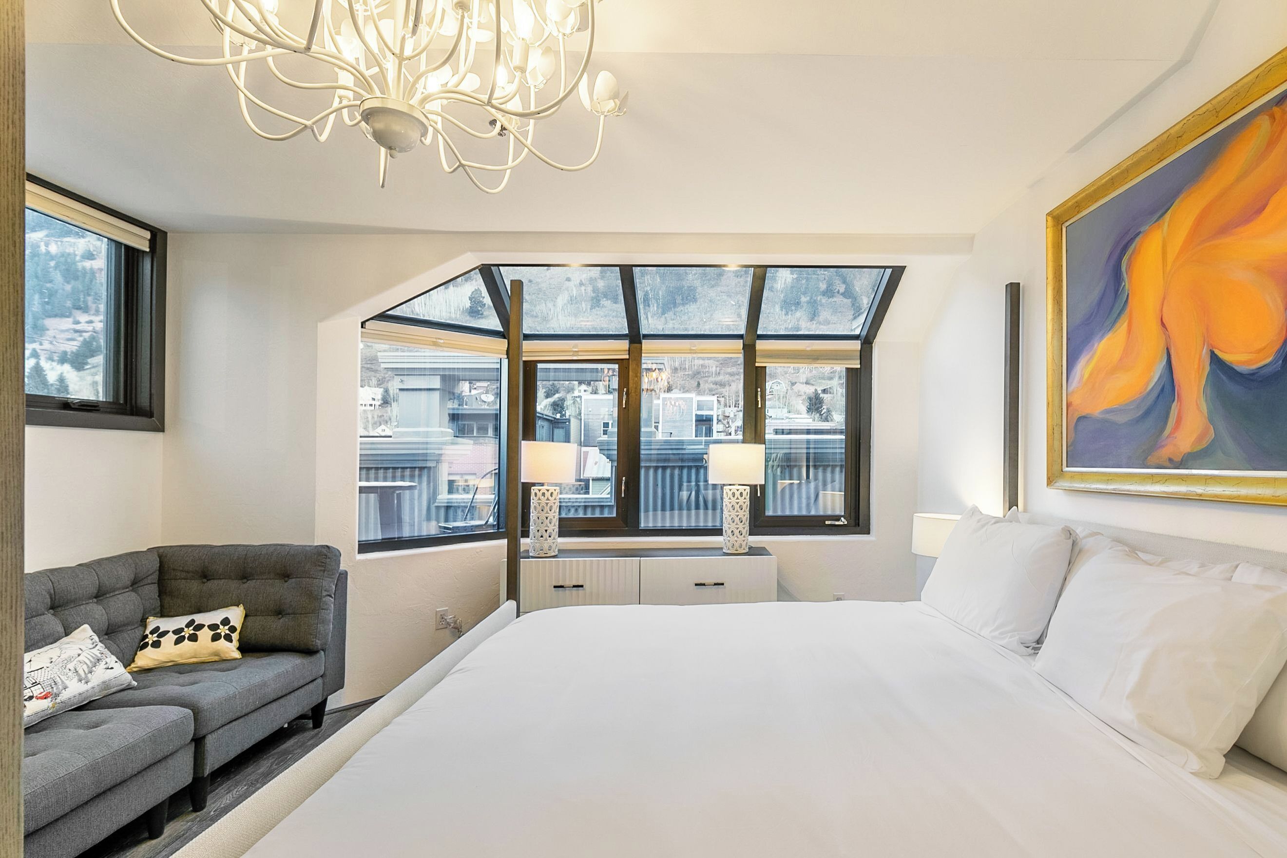 A modern hotel room with a large bed, a sofa, a chandelier, a painting, and a view of the city through large windows.