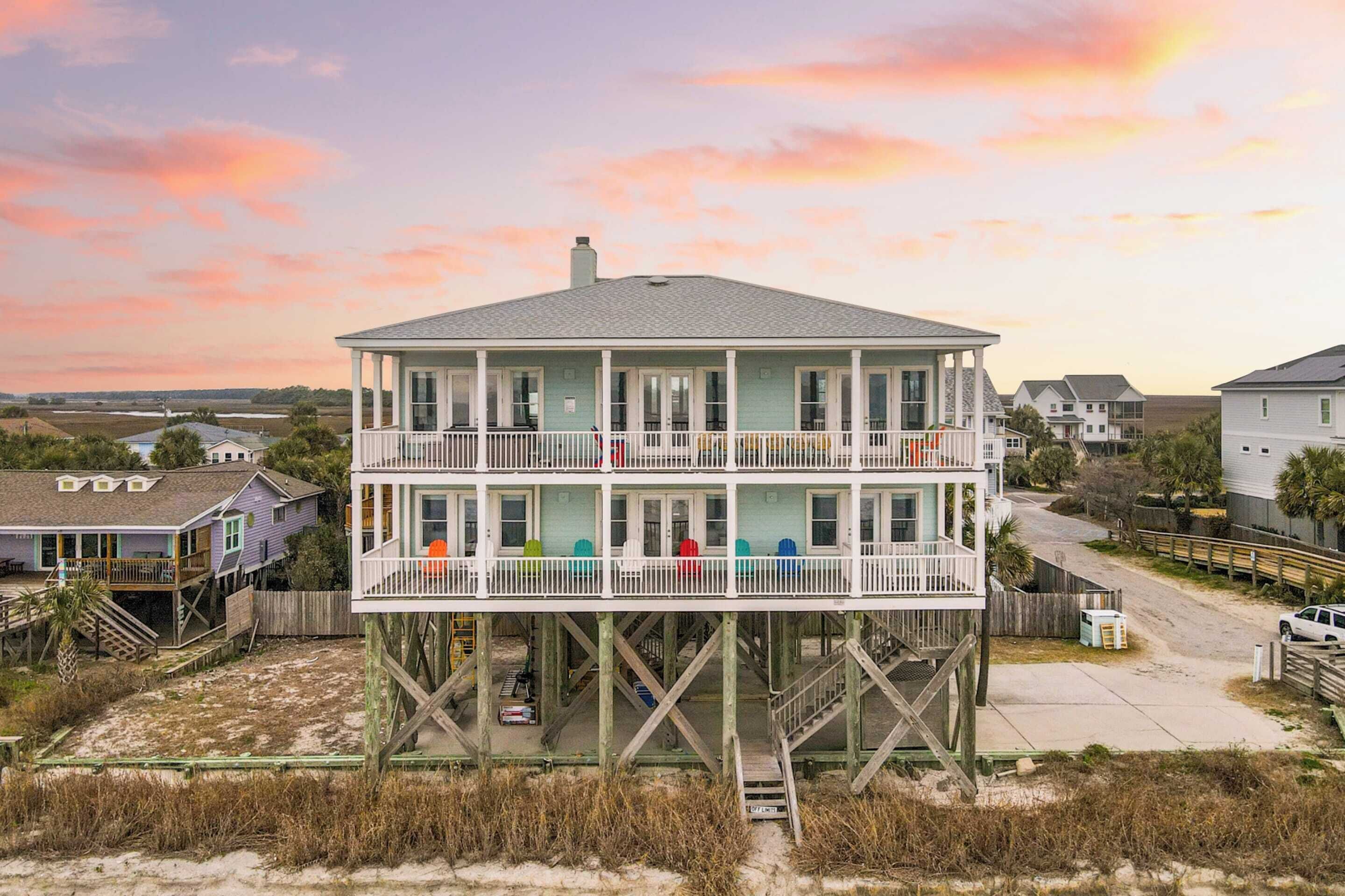 A multi-story building with a balcony on the upper floor, situated on stilts over a sandy area.