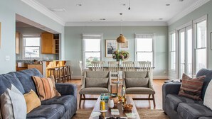 A spacious living room with a dark grey sofa, a coffee table with snacks, and a dining area with wooden chairs and a white table. - Poseidon By Avantstay On The Beach! Two Ocean Facing Balconies & Hot Tub (Folly Beach)