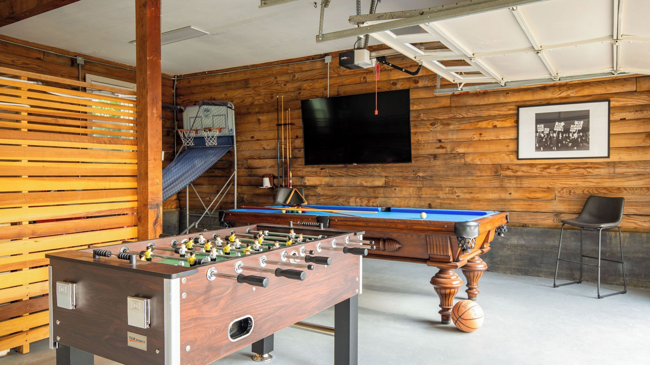 A room with a foosball table, a pool table, a television, and a framed picture on the wall.