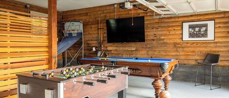 A room with a foosball table, a pool table, a television, and a framed picture on the wall.