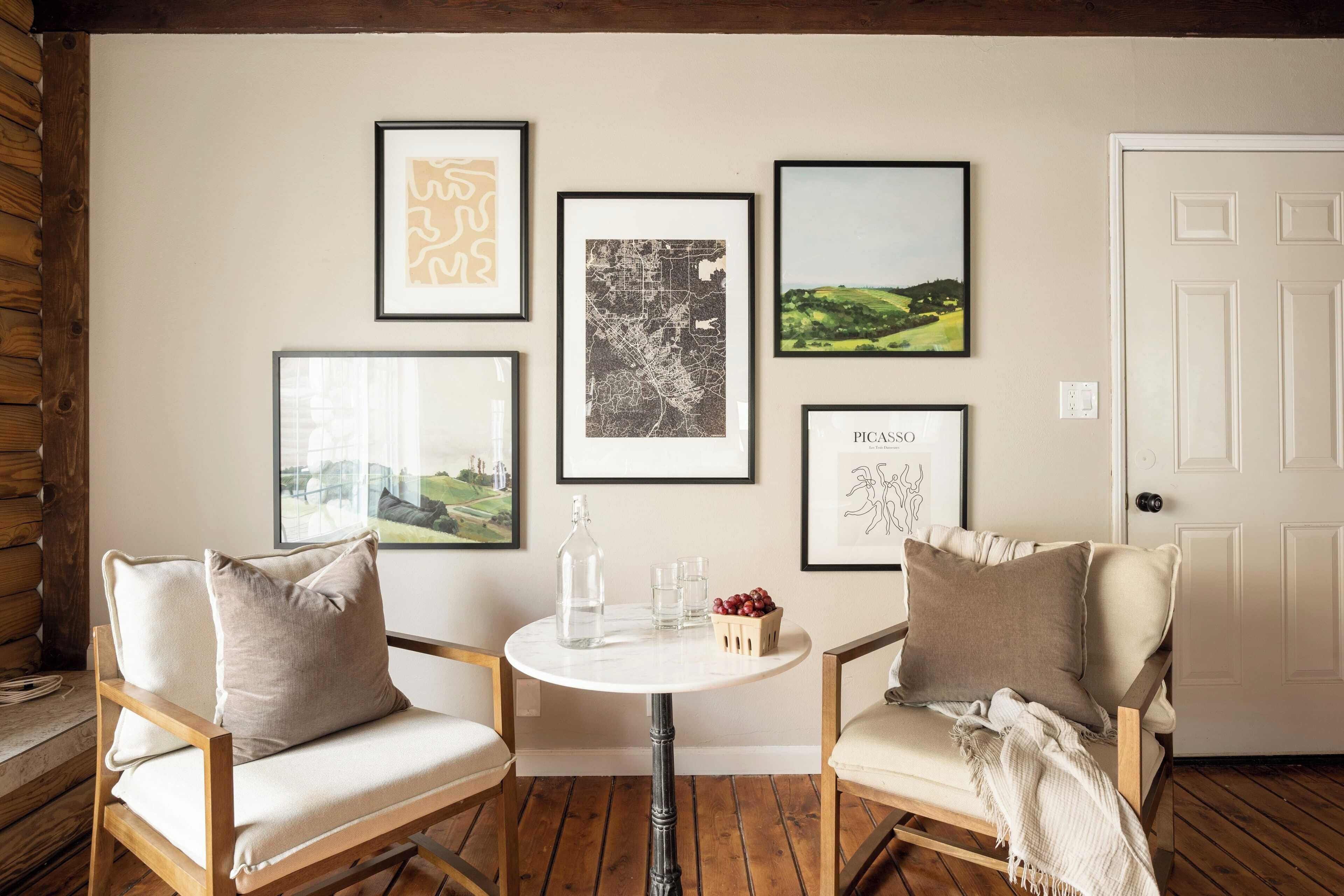 Une pièce comprenant deux chaises en bois, une table ronde avec une bouteille et des verres, et des œuvres d’art encadrées accrochées au mur.