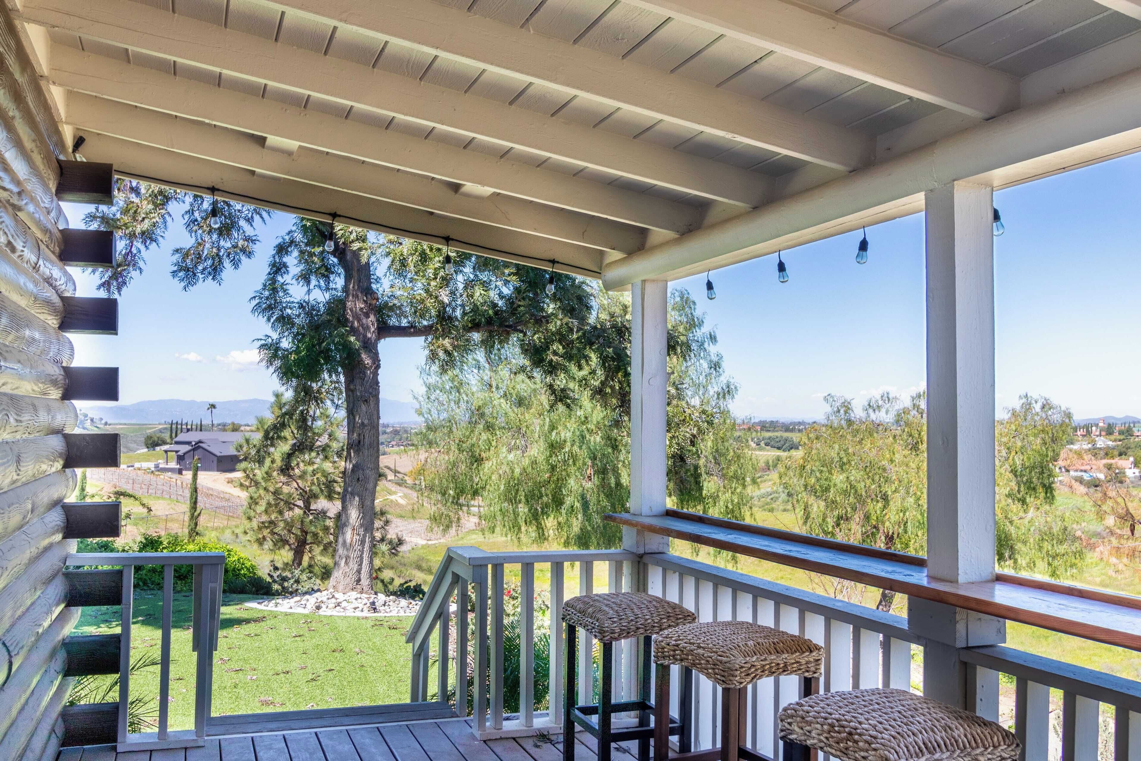 A covered porch with two wicker bar stools, a wooden deck, and a view of a grassy landscape with trees and distant buildings.