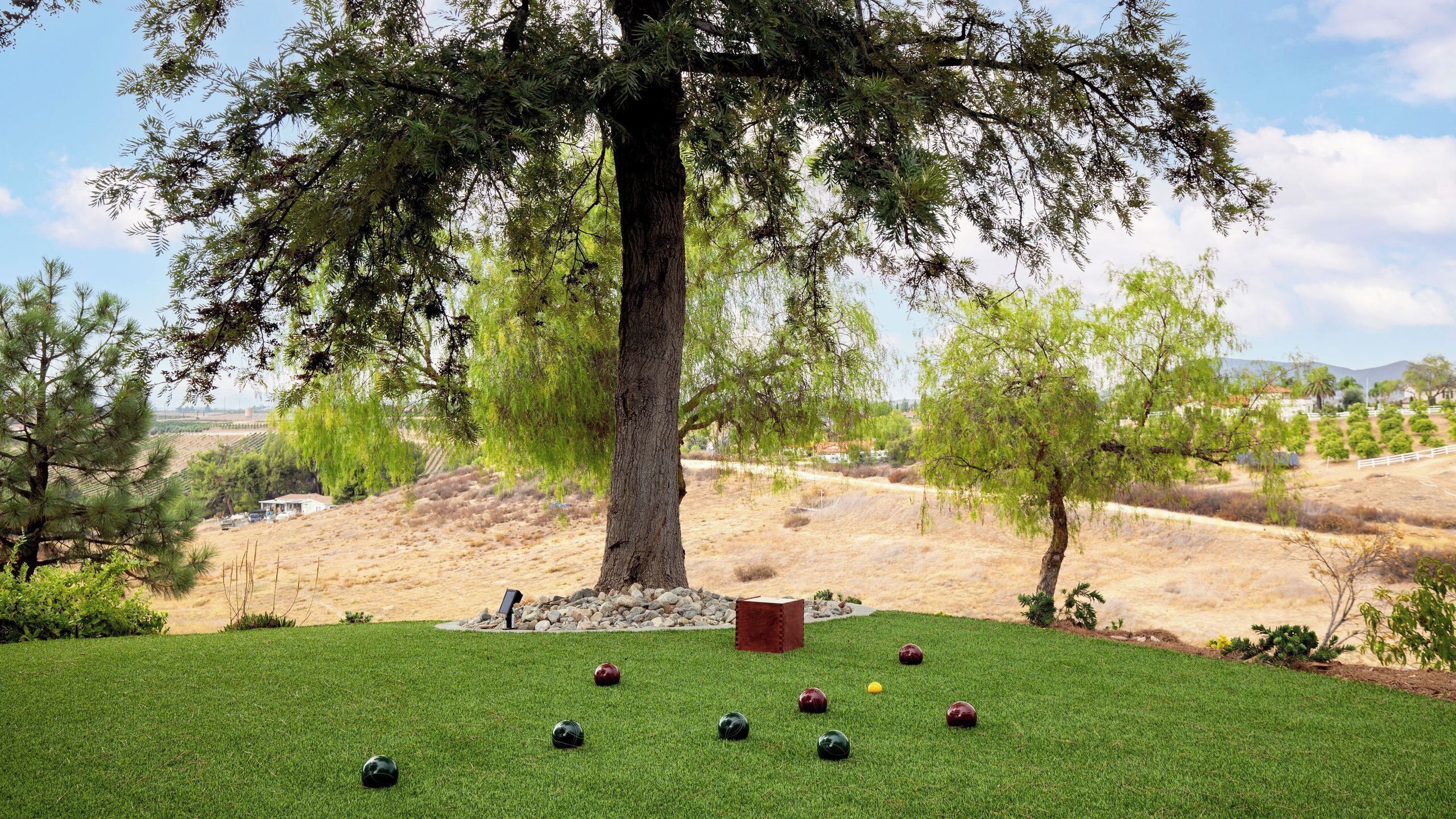 A grassy area with a large tree, a red bench, and several bocce balls.