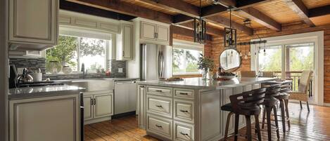 A spacious kitchen with white cabinets, a central island with bar stools, wooden flooring, and a large window providing natural light.