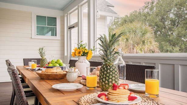A breakfast table with fruits, pancakes, and juice on a porch with a view of trees. - 1blck to Beach, Pool, Outdoor Kitchen, Game Room Pelican by Avantstay (Isle of Palms)